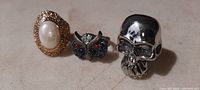 Front view of 3 costume rings showing gold-tone pearl ring, owl ring with colored stones, and silver-tone skull ring