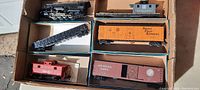 Top-down view of box containing six vintage HO scale trains including black steam locomotive, gray and red cabooses, orange and brown freight cars, and black flat car.