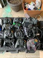 Assortment of wired video game controllers on black case including six black Xbox controllers and a purple translucent Nintendo 64 controller