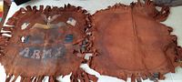 Two square leather pieces side by side, one with faded army graphics and fringed edges, the other plain with similar fringe