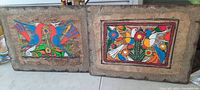 Two vintage black light glowing wall pictures side by side showing bird and floral themed artwork on solid wood backs with textured borders.