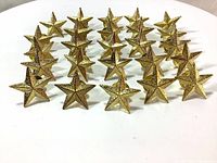Front view of 25 gold star pin badges arranged in rows on a white surface showing textured star fronts.