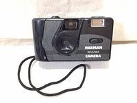 Front and top angled view of black Harman reusable 35mm point & shoot film camera showing lens, flash, wrist strap, and brand markings.