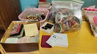 Photo showing two bags of bangles and bracelets, a basket of brooches and jewelry, and a box of boxed jewelry sets on a table.