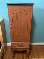 Front view of tall wooden jewelry cabinet with arched panel door and exterior drawer with brass handle.