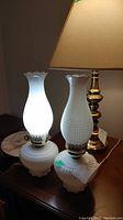 Two vintage white milk glass lamps shown lit and unlit on a wooden surface, featuring cornrow waffle pattern glass and metal collars.