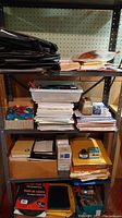 Four shelves of assorted office supplies. Top shelf contains portfolio bags and papers. Middle shelves have envelopes, writing pads, stacks of paper, plastic containers. Bottom shelf has binders, files, and boxes.