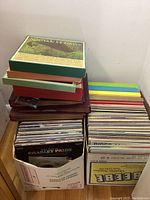 Two full white cardboard boxes of vinyl records and a stack of three boxed sets on top of a wooden surface.