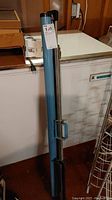 Photo of blue metal casing for projection screen with attached retractable metal pole and handle, showing screen in stored position.