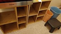Two wooden cubby shelves with six compartments each, located under a microwave, next to two trash bins and a step stool on carpet.
