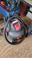 Front and side angle of gray and red Shop Vac vacuum with hose coiled around it and attachments beside it.