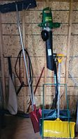 Overall view of green electric leaf blower, yellow snow shovel, red square shovel, black push broom, and dustpan with brush hanging on a wall in a workshop.