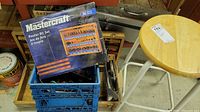 Wooden stool with light wear, Mastercraft router bit set box, metal toolbox, blue crate containing screwdrivers and tools underneath wooden cabinet.