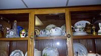 Upper shelves of wooden display cabinet showing a collection of tea cups, plates, and small decorative items including figurines.