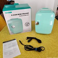 Photo of boxed Crownful mini fridge showing front of box with product image, the mini fridge unit in turquoise color, and included power cords with manual.