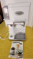 White Cuisinart Soft Serve Ice Cream Maker with control dial and dispenser handle on yellow surface with recipe booklet in front.