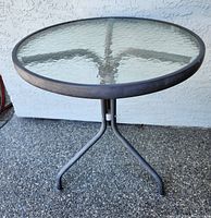 Front side view of the glass top bistro table showing round glass surface and metal frame with three legs.