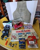 Full contents of the lot displayed on a table showing all toys and games including Monopoly board game, remote control car in packaging, tool apron, and collectible figures.