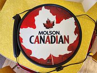 Photo of the front side of a circular Molson Canadian light-up sign with white and red leaf logo and branding.