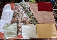 Photo showing various folded tablecloths including white embroidered, fruit and floral print, solid colors like peach and red, and light green patterned fabric.