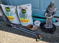 Overall image showing 2 fertilizer bags, watering hose extenders, decorative statue, and planters grouped together outside a garage door.