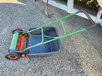 Side angled view of the Brill Luxus 38 push reel lawn mower showing green frame, black detachable grass catcher, and orange rotary blade mechanism with black wheels.