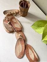 Photo showing the flat clay leaf-shaped pieces with engraved leaf patterns attached to string, alongside the cylindrical top piece, on a white surface.