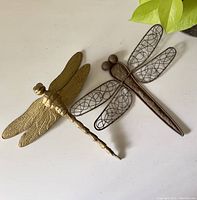 Gold-colored and dark metal dragonfly ornaments on white surface near potted plant, showing size and details.
