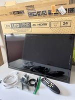 Insignia F20 24 inch Fire TV LED television with screen front view, showing accessories including remote, power cord, AV cable, and coaxial cable beside it, with original box in background.