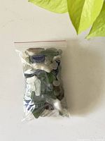 Clear plastic resealable bag filled with different colored sea glass, including green, blue, white, and clear pieces, arranged on a white surface next to a green leaf.