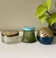 Three vintage cream jars displayed side by side on a white surface with a green plant nearby. Left jar clear with fabric floral lid, center jar green ribbed with blue embossed lid, right jar blue with gold swirl lid.