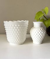 Front view of the white hobnail milk glass vase and plant holder set on a white surface with a beige background and a small green plant nearby.