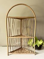 Front view of vintage small wicker corner shelf with arched top and two triangular shelves, placed near a green plant.
