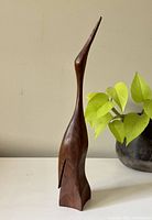 Front angle view of the vintage wooden swan sculpture next to a green leafy plant, showing its stylized flowing shape and smooth finish.