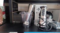Photo of Cuisinart Smart Stick wand blender with attached blending head and clear plastic measuring cup on a shelf.