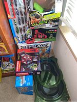 Photo showing boxed Alien Vision goggles and stunt car set, Hot Wheels cars pack, Beyblade Burst arena box, Magformers building set, and green toy racetrack arena on the floor near a window.