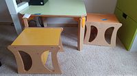 Photo of child's rectangular play table with pale green top and orange accents along with two wooden stools, one yellow topped and one orange topped, all placed on a carpet.