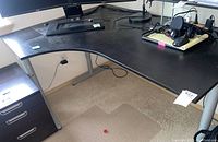 View of the IKEA Galant corner desk showing curved contour and silver metal legs, with black three-drawer filing cabinet at left.