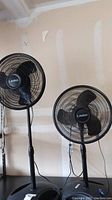 Both Lasko floor fans shown side by side against a garage wall, one taller than the other.