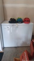 White Hotpoint chest freezer with three bicycle helmets on top, showing exterior front and surrounding area.
