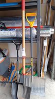 Photo showing a pointed shovel with wooden handle and black D-handle, a large metal snow shovel with yellow handle grip, and a small metal shovel with wooden handle and black D-handle standing upright.