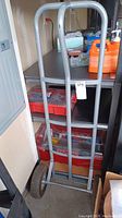 Full view of metal hand truck leaning against storage bins in a garage setting, showing frame and handle.