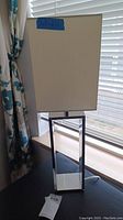 Photo of one modern table lamp featuring a white rectangular shade and chrome rectangular base on a dark wood surface