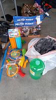 Full lot overview showing all items on concrete floor including boxed Nerf Bunkr, rackets, inflatables, water blaster, and large green jug.