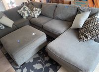 Full view of gray upholstered sectional sofa with nine pillows and matching ottoman on floral rug.