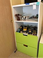 Front view of the white MDF shelving unit with two green drawers and shelves containing various toys and models.