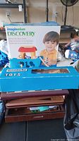 Photo shows Imaginarium Discovery Ark Shape Sorter box with child image, along with a blue box labeled Wooden Toys on top of stacked game boards.