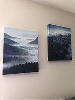 Photo showing two canvas wall art prints hanging on a white wall depicting foggy forest mountain landscape scenes with layers of trees and mist.