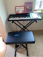 Front angled view of Yamaha PSR-E273 keyboard on black adjustable folding X-style stand with padded black bench seat in front. Three music books and black headset microphone placed on keyboard.
