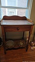 Front and side views of the antique wooden washstand placed near a window, showing the one drawer and turned legs with a scalloped back edge.